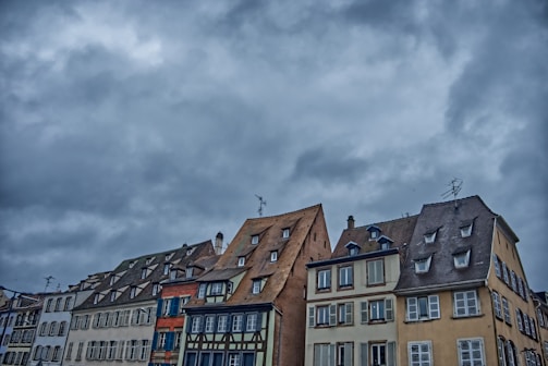 An array of traditional European buildings with steeply pitched roofs under a dramatic, cloudy sky. The architecture features a variety of colors, including cream, blue, and brown tones. Windows are prominent, with some shutters visible, adding to the historic charm.