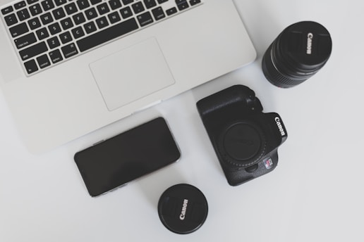 A flat lay arrangement featuring a laptop, a smartphone, a digital camera, and two camera lenses on a white surface. The laptop appears sleek and modern, while the digital camera and lenses are black and prominently display their brand.