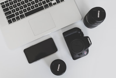 A flat lay arrangement featuring a laptop, a smartphone, a digital camera, and two camera lenses on a white surface. The laptop appears sleek and modern, while the digital camera and lenses are black and prominently display their brand.
