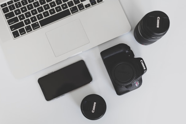 A flat lay arrangement featuring a laptop, a smartphone, a digital camera, and two camera lenses on a white surface. The laptop appears sleek and modern, while the digital camera and lenses are black and prominently display their brand.
