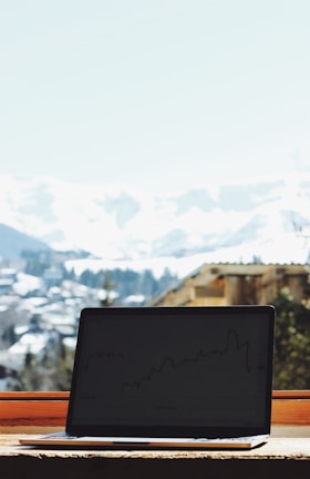 A laptop displaying a line graph sits on a wooden surface. In the background, a snowy mountain landscape and a clear blue sky are visible through a window.