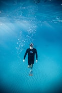 A person wearing a black wetsuit and fins is underwater, swimming in the clear blue ocean. Bubbles rise above as sunlight filters through the water, creating a serene and tranquil scene.