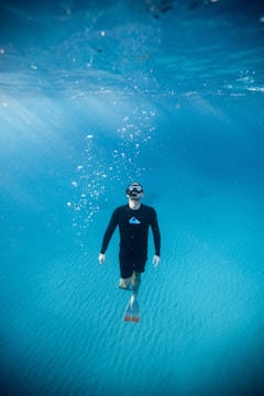A person wearing a black wetsuit and fins is underwater, swimming in the clear blue ocean. Bubbles rise above as sunlight filters through the water, creating a serene and tranquil scene.