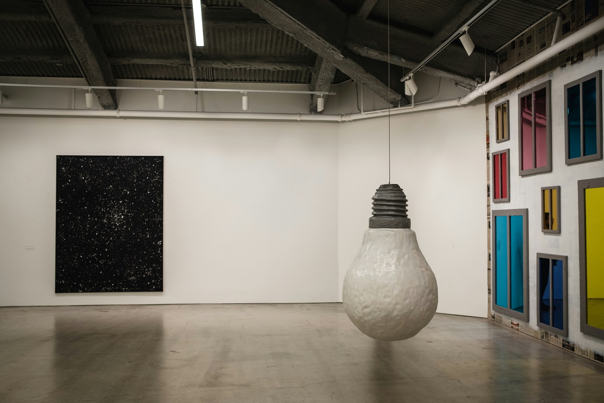 A modern art gallery featuring a large, hanging sculpture of a lightbulb in the center. The walls display a diverse collection of art pieces, including a dark, starry painting and a wall with various colorful rectangular panels resembling windows. The space has an industrial ceiling with exposed beams.