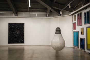 A modern art gallery featuring a large, hanging sculpture of a lightbulb in the center. The walls display a diverse collection of art pieces, including a dark, starry painting and a wall with various colorful rectangular panels resembling windows. The space has an industrial ceiling with exposed beams.