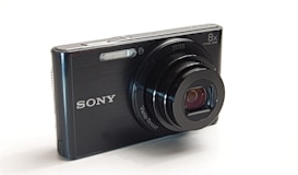 A compact digital camera with a black body and extending lens. The brand name and lens specifications are visibly printed on the camera. It features a sleek design with buttons and a flash on the top.