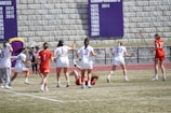 Athletes are playing a lacrosse game on a grassy field. Several players in white uniforms with lacrosse sticks are surrounding another player on the ground, who is wearing a red uniform. A stone wall in the background displays banners with championship years.