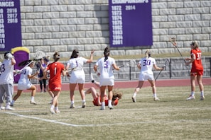 Athletes are playing a lacrosse game on a grassy field. Several players in white uniforms with lacrosse sticks are surrounding another player on the ground, who is wearing a red uniform. A stone wall in the background displays banners with championship years.
