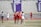 A group of female lacrosse players are on a field, with some wearing red jerseys and others white. They appear to be in the middle of a game or practice, with a referee nearby. A wall in the background features a display of men's soccer conference championship years.