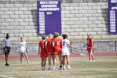 A group of female lacrosse players are on a field, with some wearing red jerseys and others white. They appear to be in the middle of a game or practice, with a referee nearby. A wall in the background features a display of men's soccer conference championship years.