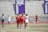 A group of female lacrosse players are on a field, with some wearing red jerseys and others white. They appear to be in the middle of a game or practice, with a referee nearby. A wall in the background features a display of men's soccer conference championship years.