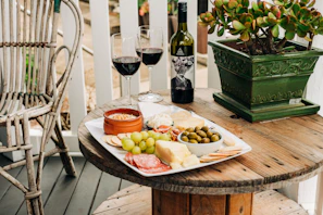 A wooden spool table set on a patio holds a charcuterie board with a variety of cheeses, crackers, salami, grapes, and olives. Two glasses of red wine and a bottle of wine accompany the board. A green, leafy plant in a decorative pot adds a touch of nature. A wicker chair is positioned nearby.