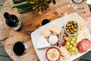 Olives and other snacks on white ceramic plate beside wine glass and bottle