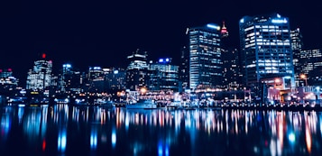 A vibrant cityscape at night with skyscrapers lit up, reflecting brilliantly in a body of water. The image captures the urban skyline with bright lights and a bustling ambiance.