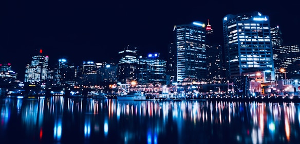 A vibrant cityscape at night with skyscrapers lit up, reflecting brilliantly in a body of water. The image captures the urban skyline with bright lights and a bustling ambiance.