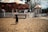 A young child is walking on a dirt ground toward a colorful carousel with animal figures, surrounded by a white picket fence. There is a small ticket booth near the carousel displaying a sign with prices, and people are visible enjoying the ride. The scene is bright and sunny with trees in the background.