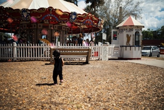 A young child is walking on a dirt ground toward a colorful carousel with animal figures, surrounded by a white picket fence. There is a small ticket booth near the carousel displaying a sign with prices, and people are visible enjoying the ride. The scene is bright and sunny with trees in the background.