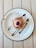 A dessert is placed on a white plate with a black rim, topped with pink frosting and sprinkled with chocolate crumbs. Chocolate sauce is artfully drizzled on the plate, complementing the dessert's presentation. The plate rests on a light-colored wooden table.