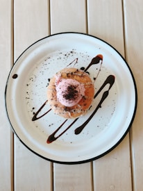 A dessert is placed on a white plate with a black rim, topped with pink frosting and sprinkled with chocolate crumbs. Chocolate sauce is artfully drizzled on the plate, complementing the dessert's presentation. The plate rests on a light-colored wooden table.