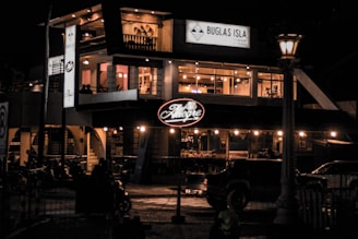 A two-story building with prominent signage illuminated at night. The upper floor has large glass windows showcasing an inviting interior with warm lighting and decor. The establishment appears to be a restaurant or bar, and there are vehicles and lampposts visible outside, adding to the urban nighttime setting.