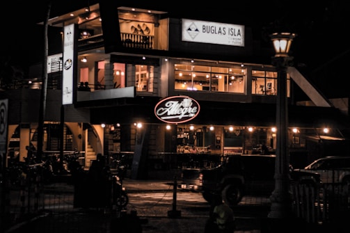 A two-story building with prominent signage illuminated at night. The upper floor has large glass windows showcasing an inviting interior with warm lighting and decor. The establishment appears to be a restaurant or bar, and there are vehicles and lampposts visible outside, adding to the urban nighttime setting.
