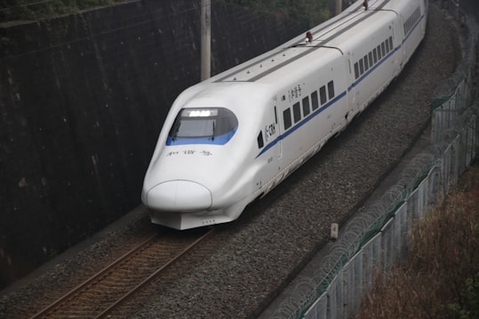 A sleek, white bullet train is captured speeding along a set of tracks, situated in a narrow passage with a tall retaining wall on one side and a metal fence on the other. The train's streamlined design and large windows are prominent.