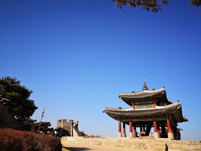 A traditional Korean pavilion with intricate roof decorations and red pillars stands prominently. To the left, an ancient stone wall with battlements is visible, accompanied by a flagpole with a flag. The sky is clear and blue, and trees with dense foliage frame the scene.