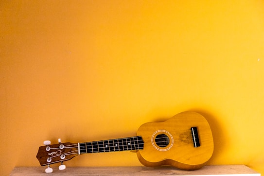 A ukulele lies on a light wooden surface against a vibrant yellow wall. The ukulele's body and neck are prominently visible, with tuning pegs and strings clearly defined.