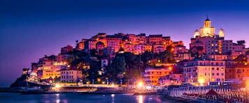 A picturesque coastal town illuminated at night, featuring a cluster of colorful buildings ascending a hill. The warm glow from streetlights and windows contrasts with the deep blue of the evening sky. A prominent church with a lit dome crowns the hill, adding an architectural focal point to the scene. Below, beach umbrellas are aligned along the shoreline, hinting at a seaside location.
