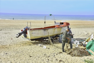 A boat resting on sandy beach beside the ocean, with two individuals working on fishing nets nearby. The ocean stretches into the horizon, and a few scattered items are visible on the sand.