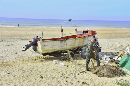 A boat resting on sandy beach beside the ocean, with two individuals working on fishing nets nearby. The ocean stretches into the horizon, and a few scattered items are visible on the sand.