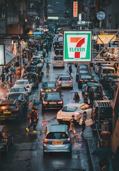 A busy city street scene with heavy traffic and pedestrians carrying umbrellas. The area is wet, possibly due to recent rain. Brightly lit signs, including a prominent 7-Eleven sign, are visible among various shops and buildings. A diverse array of cars and people can be seen navigating through the bustling environment.