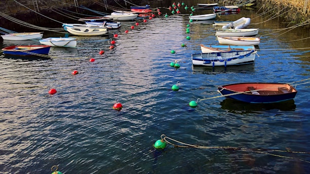 A group of small boats are moored in a calm harbor, surrounded by vibrant green and red buoys. The boats vary in color and size, and they are tethered with ropes to the edge of the stone-lined dock.
