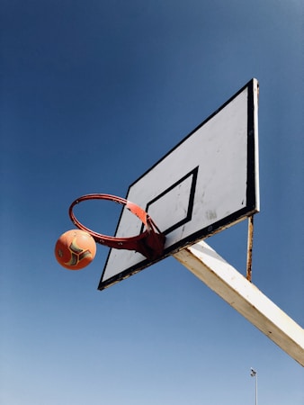 A basketball is captured mid-air, about to pass through a slightly tilted red hoop attached to a weathered white backboard. The sky forms a clear blue background, enhancing the vividness of the scene.