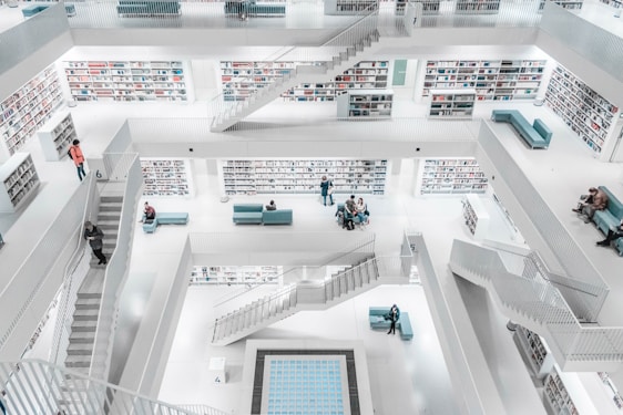 A spacious, modern library with multiple levels and open balconies. The bright white environment is contrasted by teal-colored couches and staircases connecting the floors. Rows of bookshelves line the walls, and a few people are visible, some walking, others sitting and reading or talking.