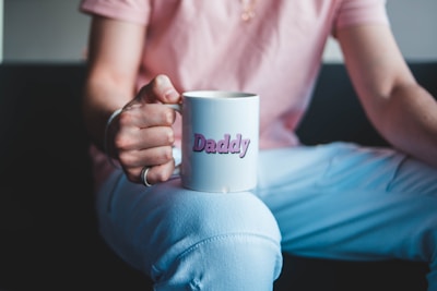 A person is sitting, holding a white mug with the word 'Daddy' printed in pink. The individual is wearing a pink shirt and white pants, with the mug resting on their knee.