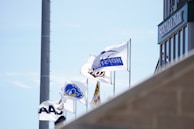 Flags are waving in the wind against a clear blue sky, adjacent to the corner of a building with 'SENTAKA PARK' signage. The flags have various logos and designs, representing different organizations and teams.