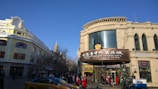 A bustling street scene in a city featuring a large, ornate building with signage in both English and another language. The building is labeled 'Andrew Foreign Goods City.' It has a classic architectural style with large windows and decorative details. Pedestrians, including people of various ages and a taxi, are visible. The sky is clear and blue.