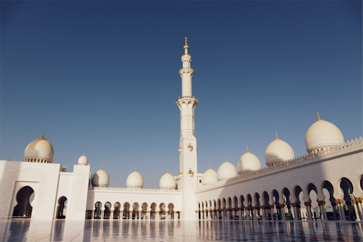 An expansive courtyard features majestic white architecture with large domes and a tall minaret. The design is intricate, reflecting traditional Islamic style. The clear blue sky enhances the serene and grand ambiance.