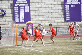 Athletes are actively engaged in a competitive lacrosse game on a field. Players in red uniforms and players in white uniforms are interacting near a goal post. A referee is present, and banners displaying championship years are visible in the background.