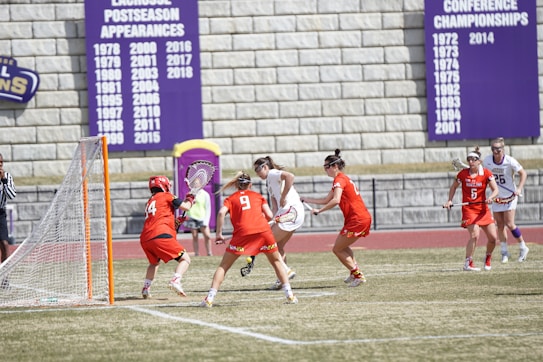 Athletes are actively engaged in a competitive lacrosse game on a field. Players in red uniforms and players in white uniforms are interacting near a goal post. A referee is present, and banners displaying championship years are visible in the background.