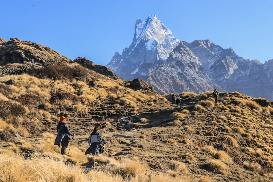 The image depicts a group of hikers ascending a mountain trail, with a majestic snow-capped peak visible in the background. The hikers are dressed in outdoor gear and carrying backpacks, suggesting they are well-prepared for their journey. The perspective is from ground level, looking up at the hikers as they make their way towards the top of the mountain.