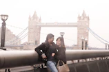 A professional couple walking near London’s Tower Bridge, holding passports and visas with confident smiles.