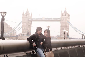 A professional couple walking near London’s Tower Bridge, holding passports and visas with confident smiles.