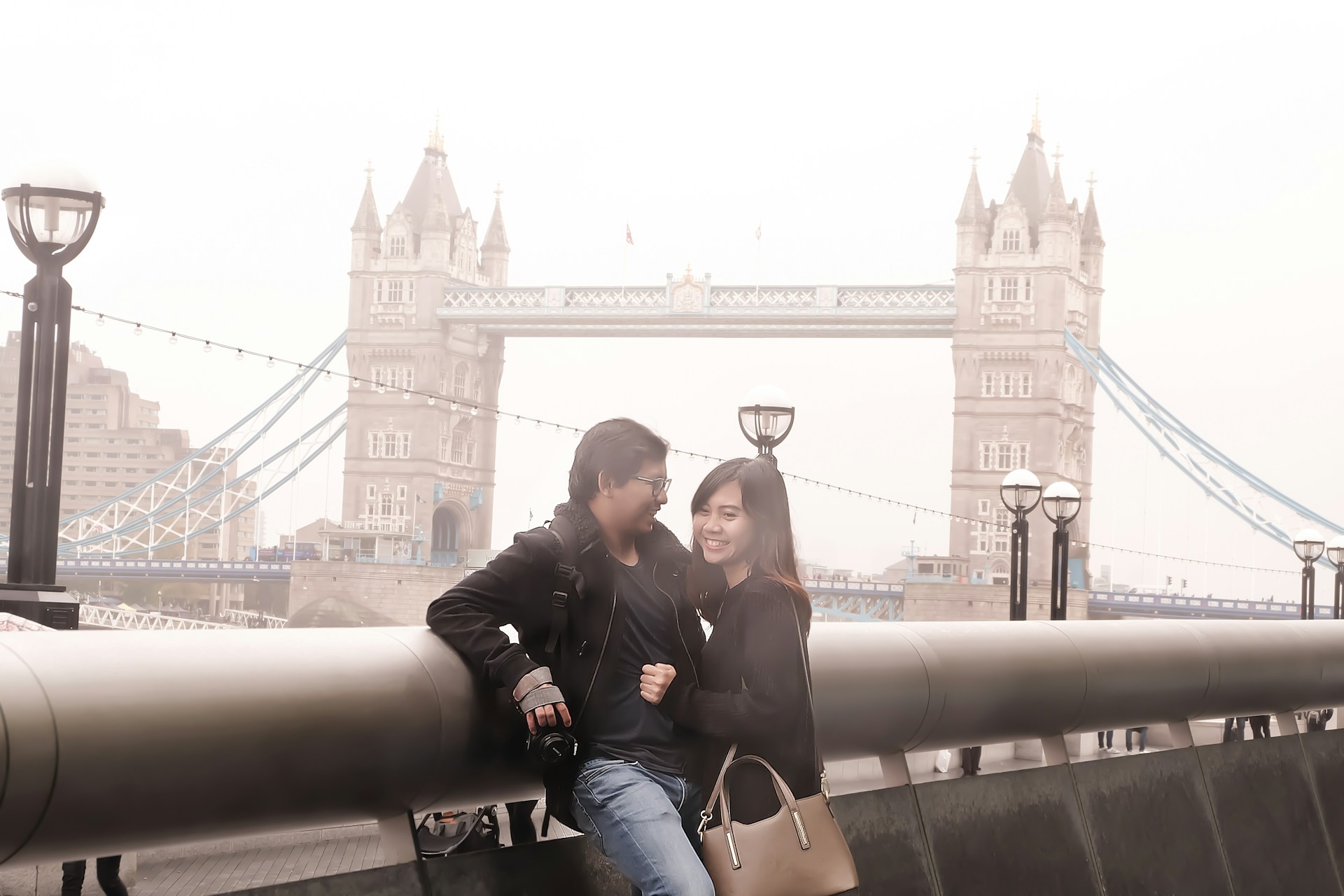 A couple is posing and smiling while leaning against a railing with Tower Bridge in the background. The setting appears foggy, and there are a few street lamps lining the walkway. The man is holding a camera and wearing casual clothing with a jacket, and the woman is carrying a handbag.