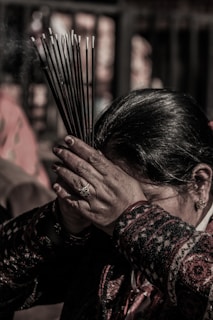 A person holds a bundle of incense sticks close to their forehead, with both hands placed together in a gesture of reverence or prayer. The background is blurred, and the person is wearing a patterned garment.