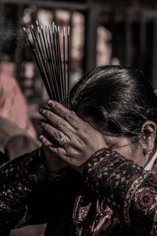 A person holds a bundle of incense sticks close to their forehead, with both hands placed together in a gesture of reverence or prayer. The background is blurred, and the person is wearing a patterned garment.