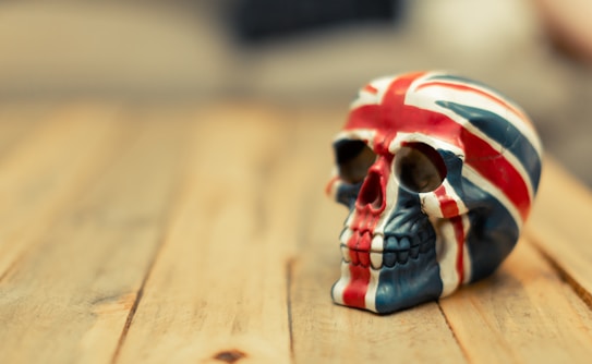 A skull sculpture with the design of the United Kingdom's Union Jack flag sits on a wooden surface. The skull is painted in red, white, and blue, with the flag pattern covering the entire surface. The background is blurred, emphasizing the detailed artwork on the skull.
