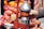 A hand-operated juicer is squeezing a pomegranate half, surrounded by other halved and whole pomegranates. The juicer is metallic with a red fixture, and the environment resembles a fruit market or street vendor setting. The scene includes vivid red and orange hues.