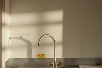 A minimalist kitchen scene featuring a sleek metallic faucet above a sink. Soft shadow patterns of a window are cast on the wall, creating a tranquil ambiance. A small yellow warning sign indicating 'very hot water' is visible near the faucet.